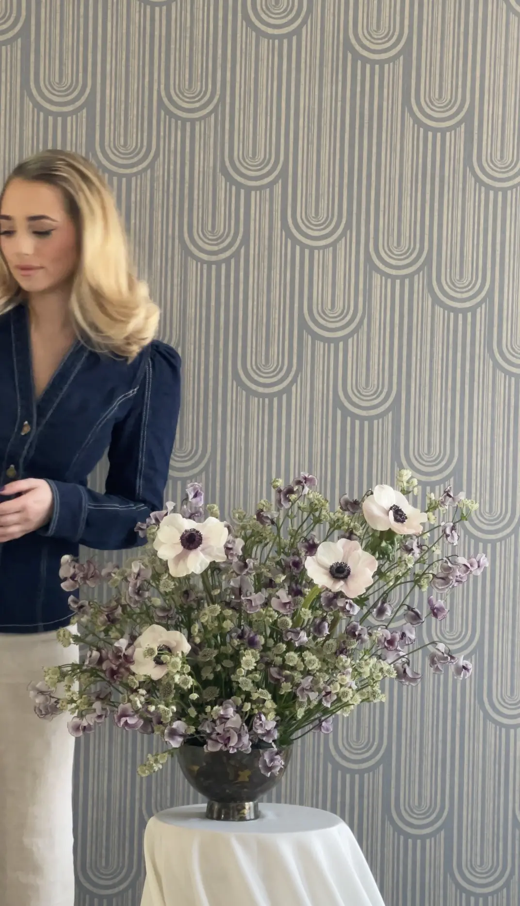 Woman in a dark blazer arranging flowers against a patterned wall.